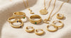 A luxury collection of Cheriva Jewels solid gold fine jewelry, featuring delicate pendant necklaces, minimalist hoop earrings, and stacked gold rings set with green and blue gemstones on a soft beige fabric background.